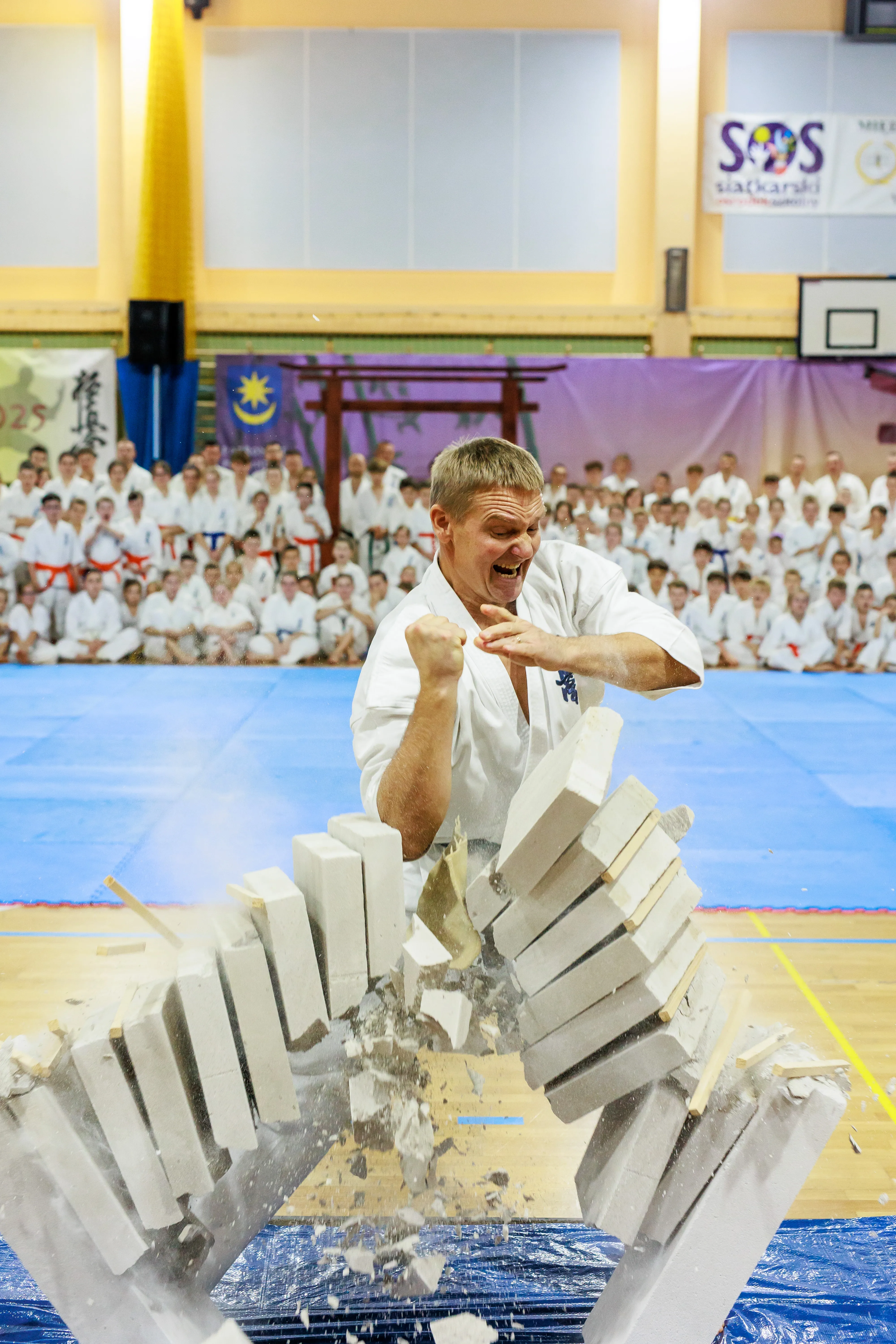 Mężczyzna w białym kimono uderza pięścią w stos białych cegieł łamiąc je na dwie części na hali sportowej, w tle widoczna grupa osób w białych strojach martial arts siedzących na niebieskiej macie.
