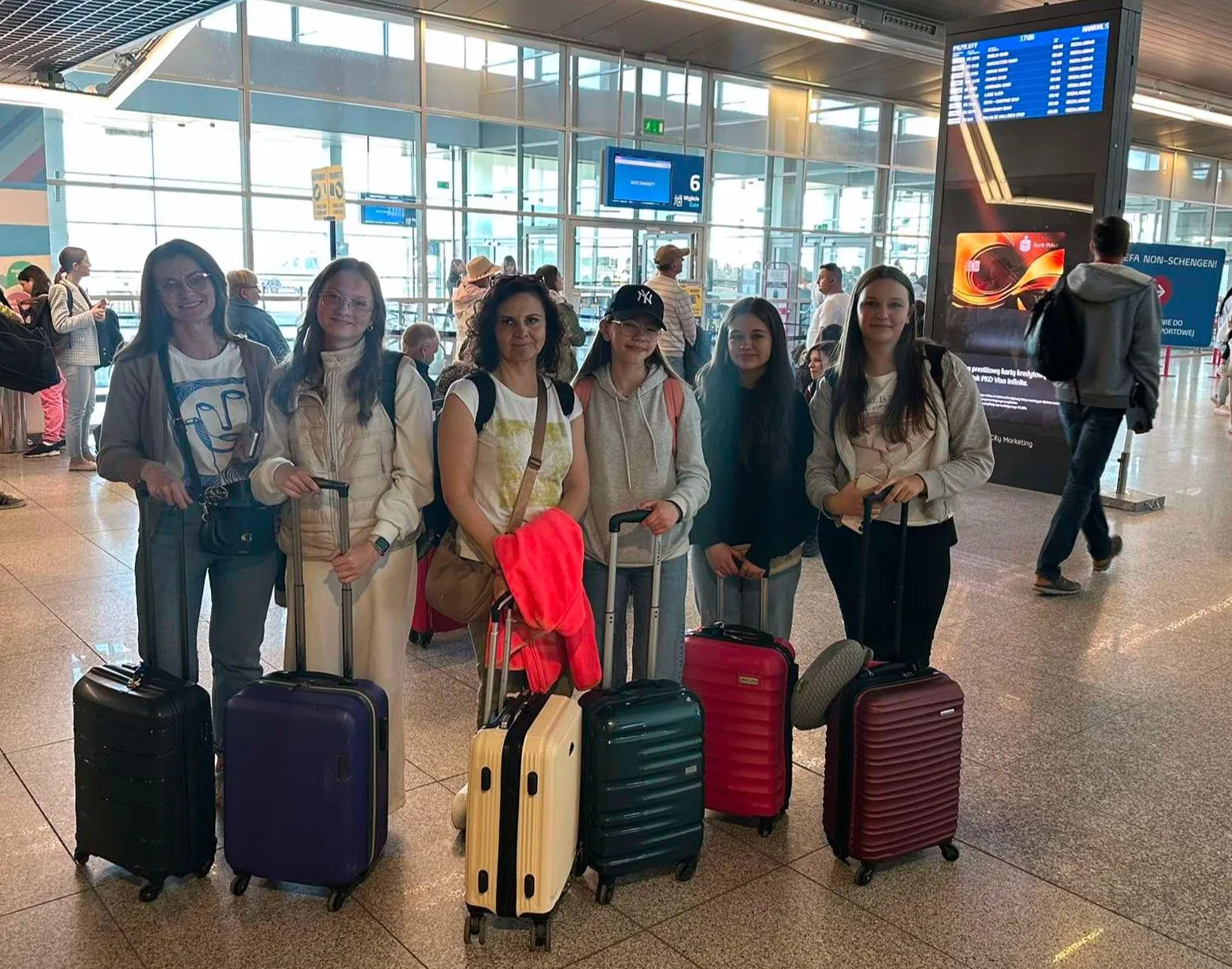 Sześć kobiet stoi w hali lotniska z walizkami na kółkach. W tle widać szklane ściany, tablice informacyjne oraz inne osoby. Scena rozgrywa się w ciągu dnia.