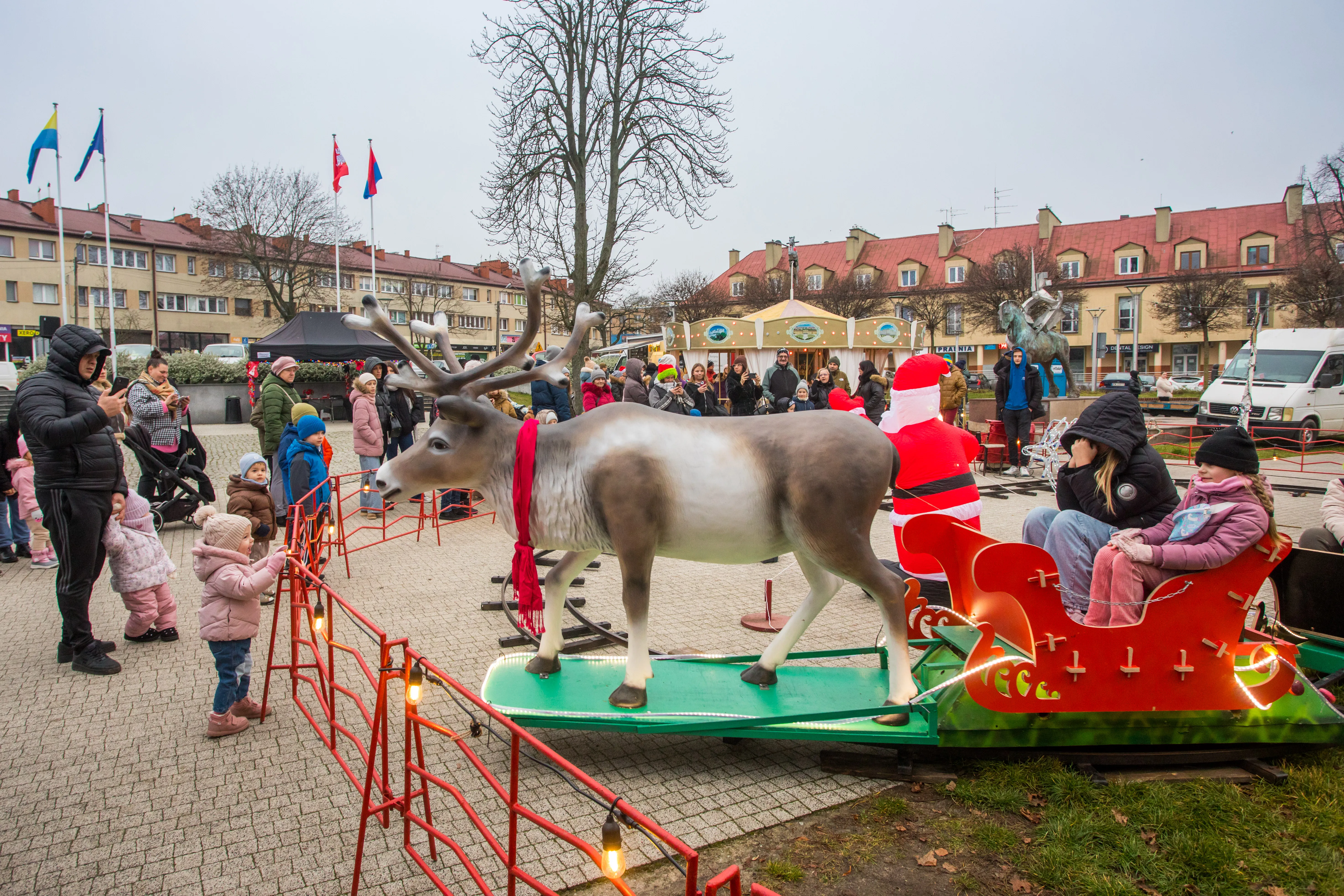 Plac miejski z modelem renifera i saniami św. Mikołaja, wokół ogrodzenie z lampkami, na saniach siedzą dwie osoby. Ludzie w zimowych ubraniach stoją i fotografują atrakcję, w tle budynki mieszkalne i karuzela. Dzień, pochmurno.