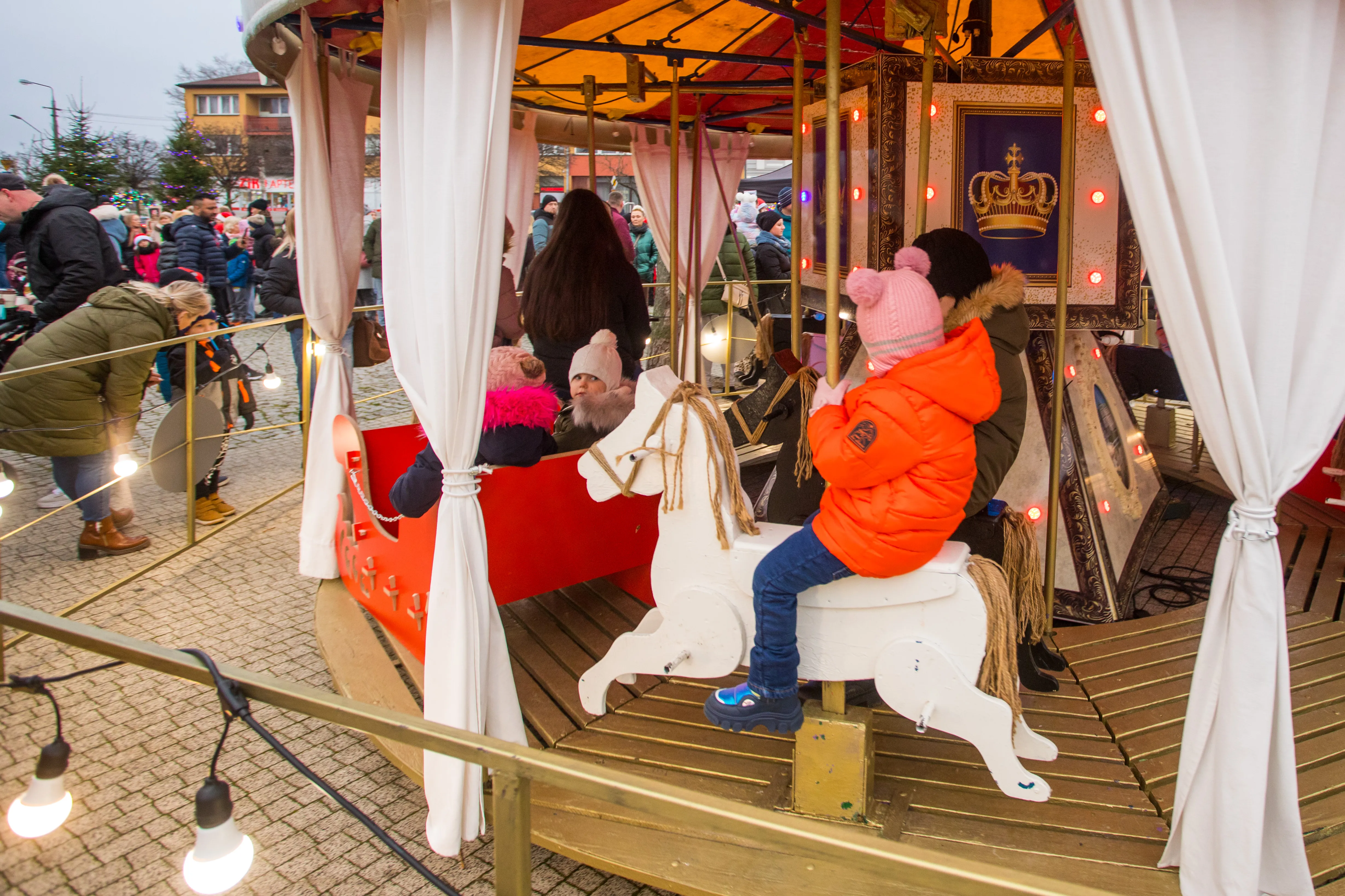 Dzieci bawiące się na karuzeli z drewnianymi konikami i czerwonym wózkiem. Karuzela znajduje się na zewnątrz, wśród zgromadzonych osób ubranych na zimę. W tle budynki i ozdobione światełkami drzewo. Wieczorna pora.