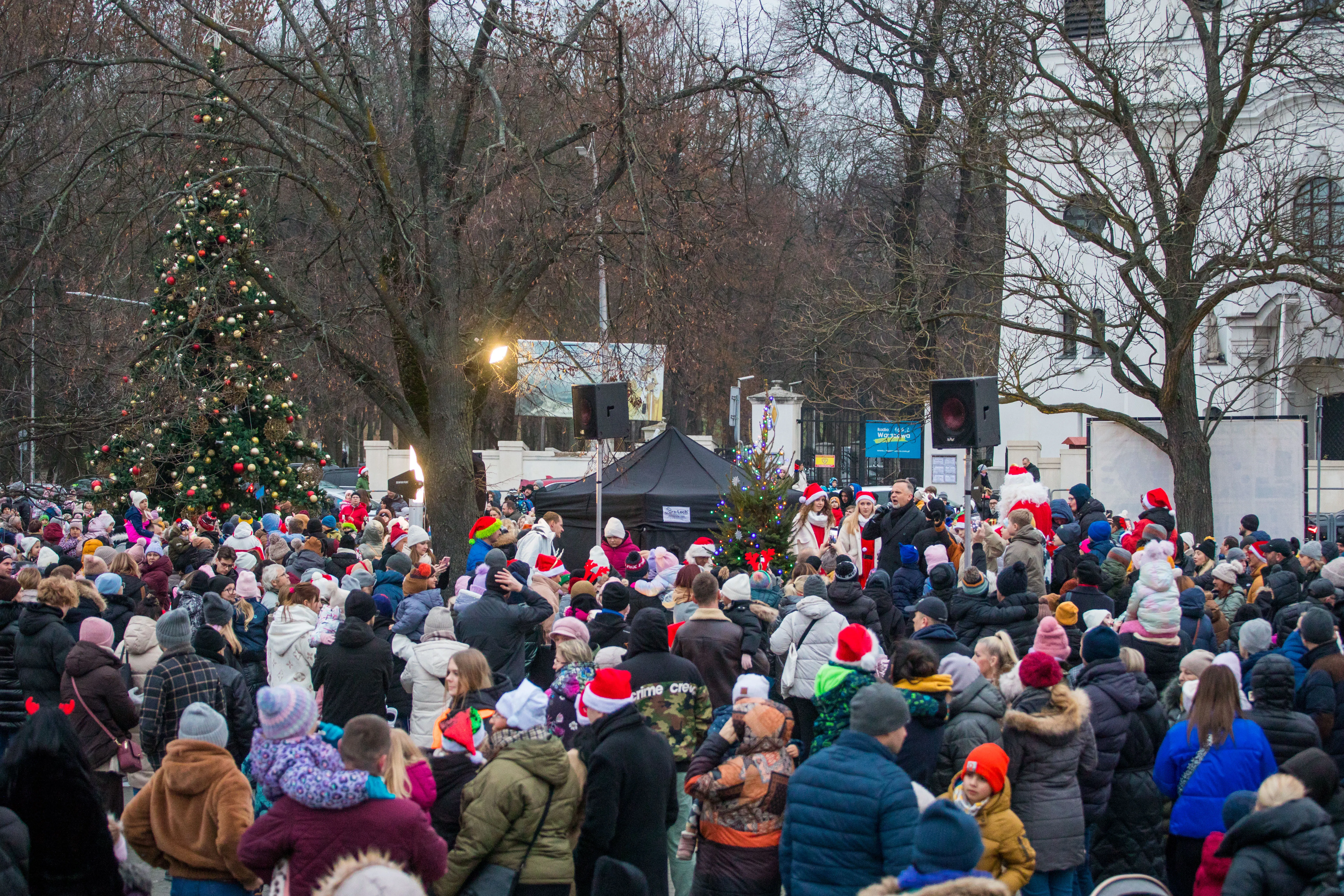 Duża grupa ludzi zgromadzona na zewnątrz w okresie zimowym, wiele osób nosi czapki świąteczne typu Mikołaj, w tle dwa przystrojone choinki i nagłośnienie przy namiocie.