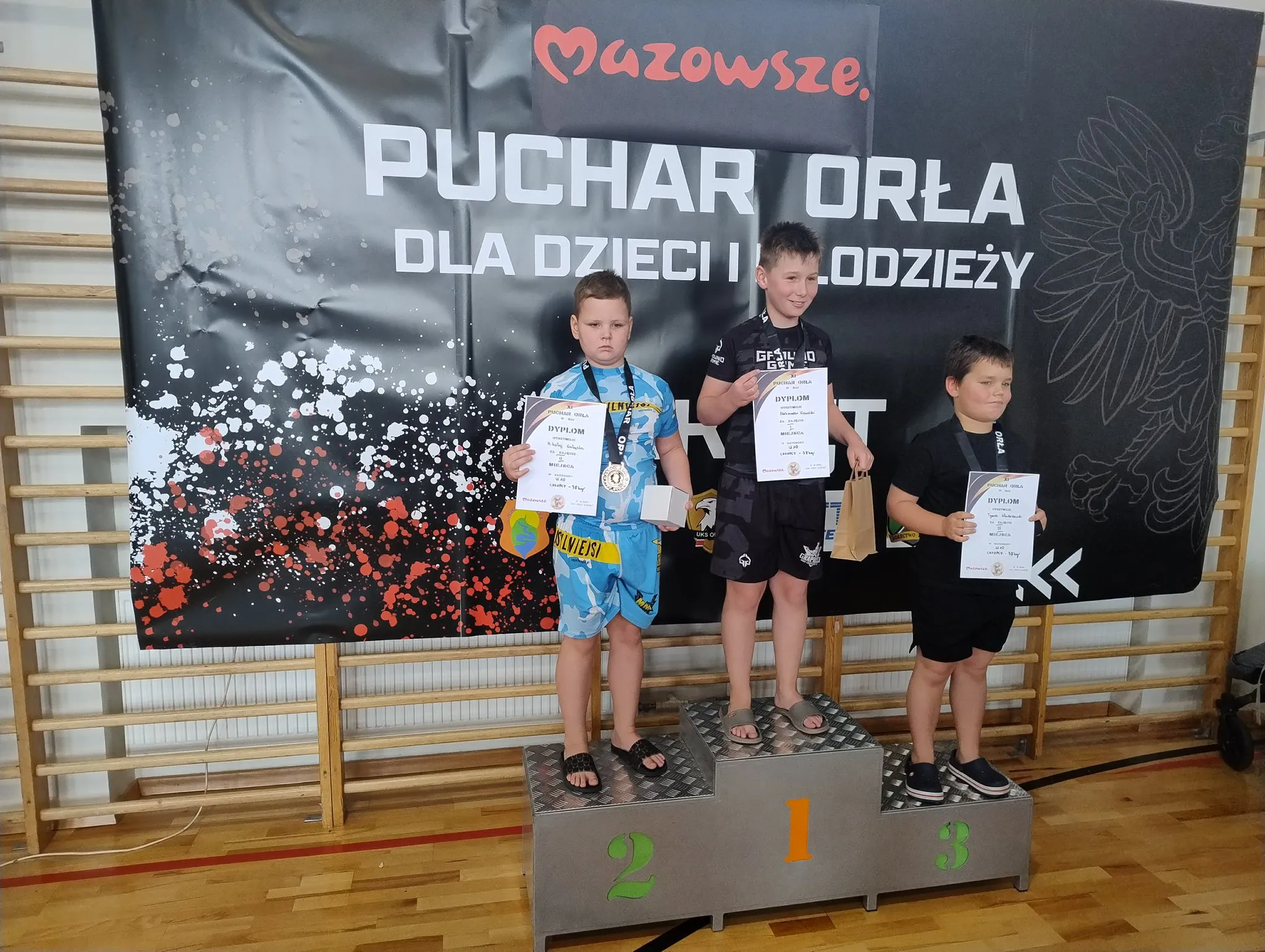 Trójka chłopców stoi na podium w hali sportowej. Chłopiec na pierwszym miejscu ma czarne ubranie i trzyma dyplom oraz torebkę. Chłopiec na drugim miejscu nosi niebiesko-żółty strój sportowy i trzyma dyplom. Chłopiec na trzecim miejscu ma czarny strój i trzyma dyplom. Za nimi czarna ścianka z napisem „Puchar Orła dla dzieci i młodzieży” oraz „Mazowsze”.