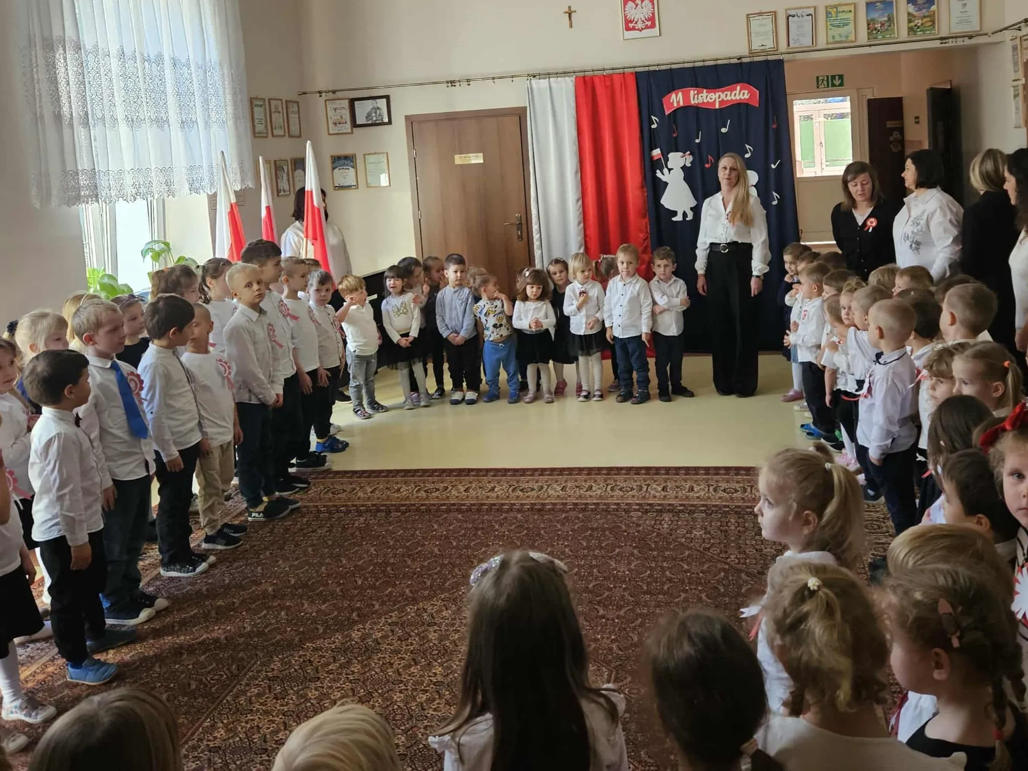 Grupa dzieci i kilka dorosłych stoi w półkolu w sali z jasnymi ścianami i dużym oknem z białą firanką. W tle polskie flagi oraz dekoracja z napisem „11 listopada” na czerwono-biało-granatowym tle. Na podłodze znajduje się brązowy dywan.
