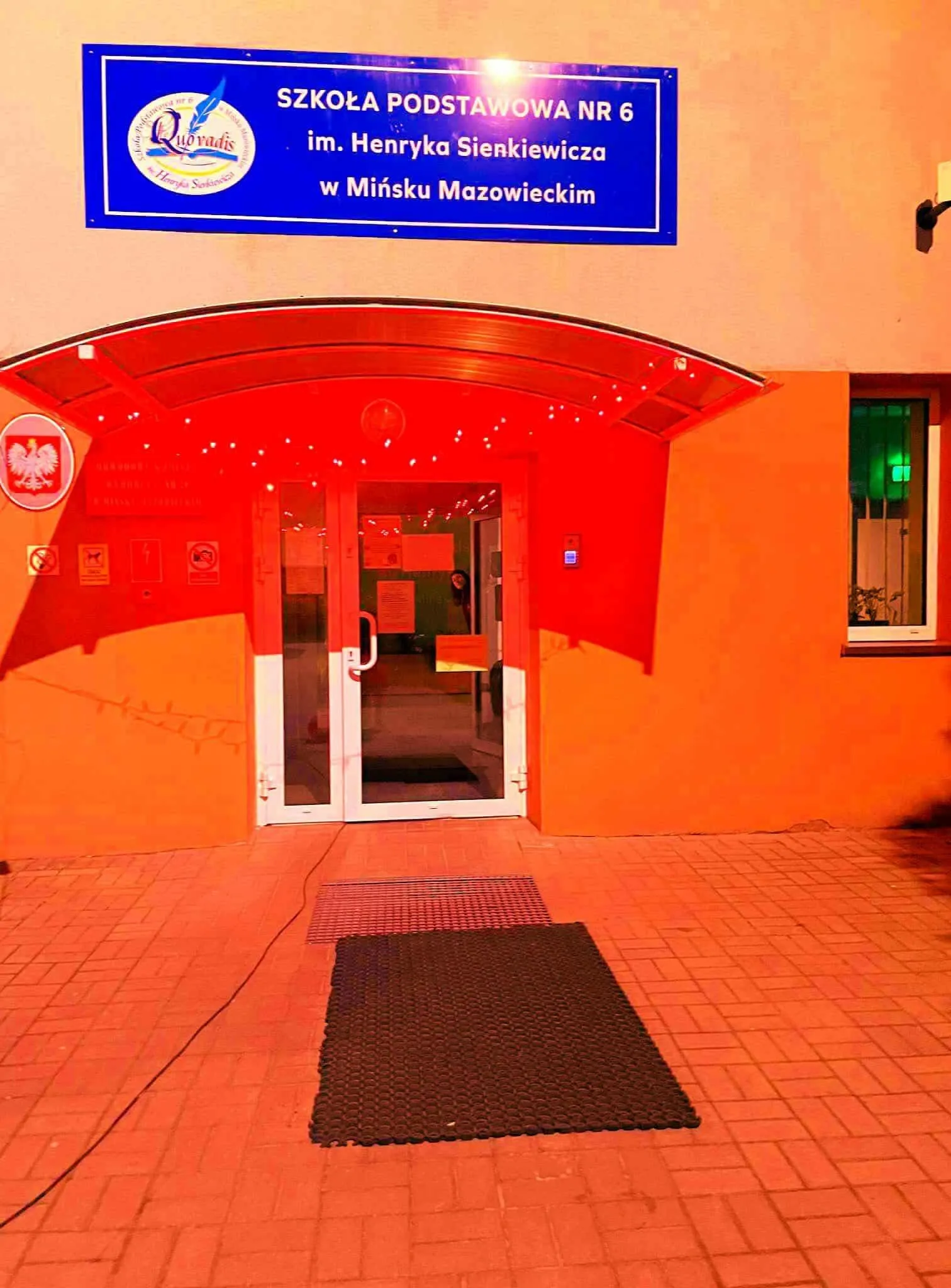 Wejście do budynku szkoły podstawowej z białymi drzwiami, nad którym znajduje się niebieska tablica z nazwą szkoły na pomarańczowym tle. Po prawej stronie okno, a przed wejściem położona jest czarna mata na czerwonej kostce brukowej.