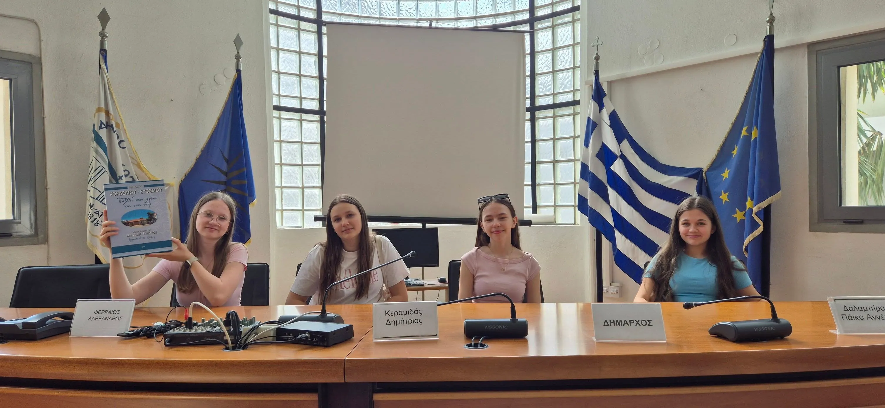 Cztery młode kobiety siedzą przy długim stole konferencyjnym w jasnym pomieszczeniu z białymi ścianami. Za nimi wiszą trzy flagi oraz stoi ekran projekcyjny. Na stole znajdują się mikrofony i tabliczki z imionami.