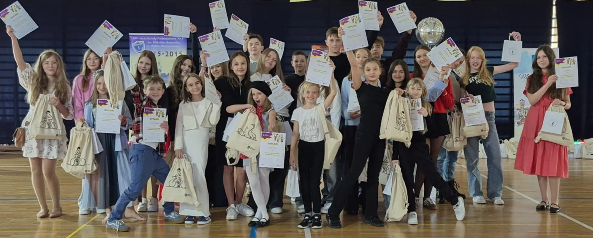 Grupa dzieci i nastolatków stoi na hali sportowej, trzymając w górze dyplomy i torby z nadrukiem roweru. W tle widoczna jest tablica informacyjna i ściana z ciemnym tłem.