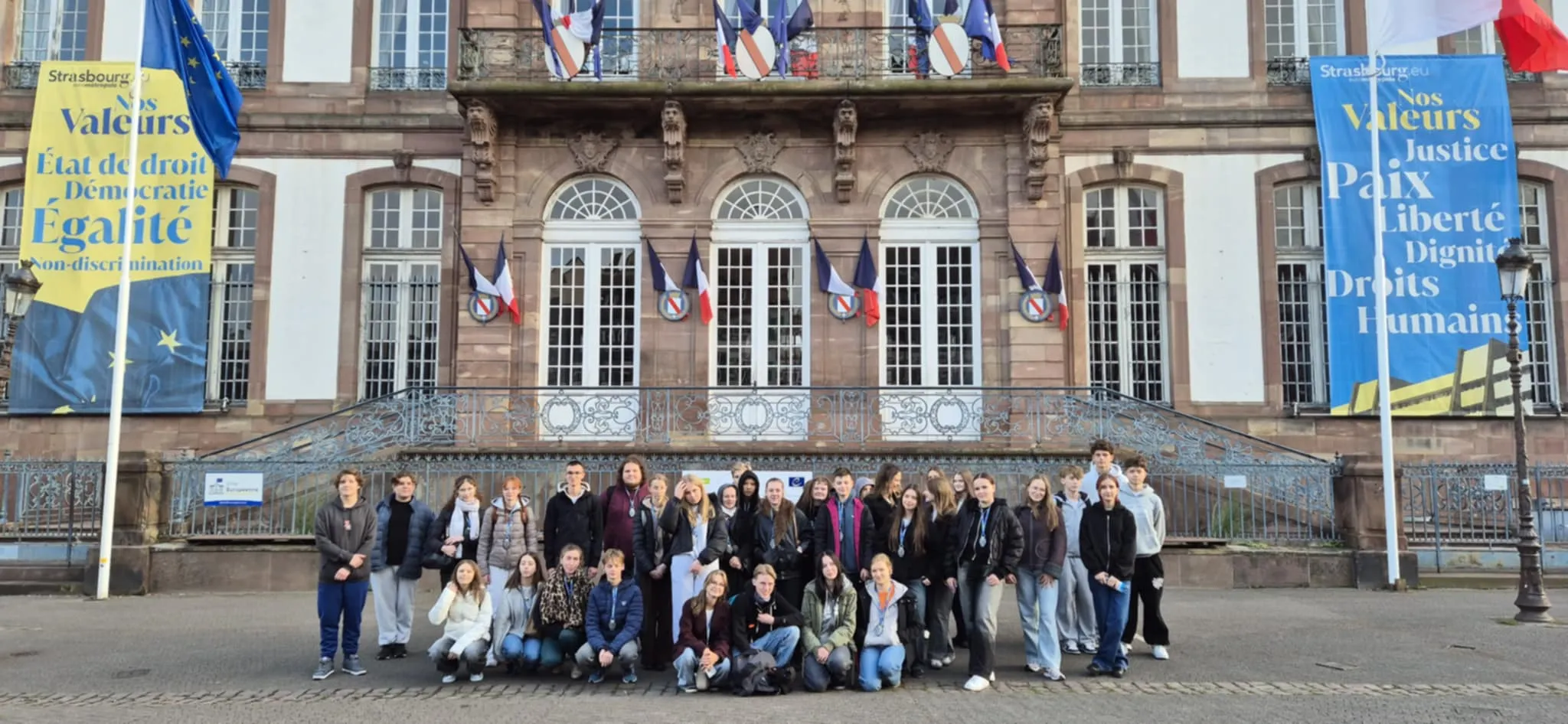 Grupa około 30 osób stoi i kuca na chodniku przed dużym budynkiem z kilkoma wysokimi, białymi oknami. Budynek jest ozdobiony flagami Francji oraz dużymi banerami w kolorach żółtym i niebieskim po obu stronach wejścia. Na banerach widoczne są napisy w języku francuskim. Dzień, miejsce na zewnątrz.