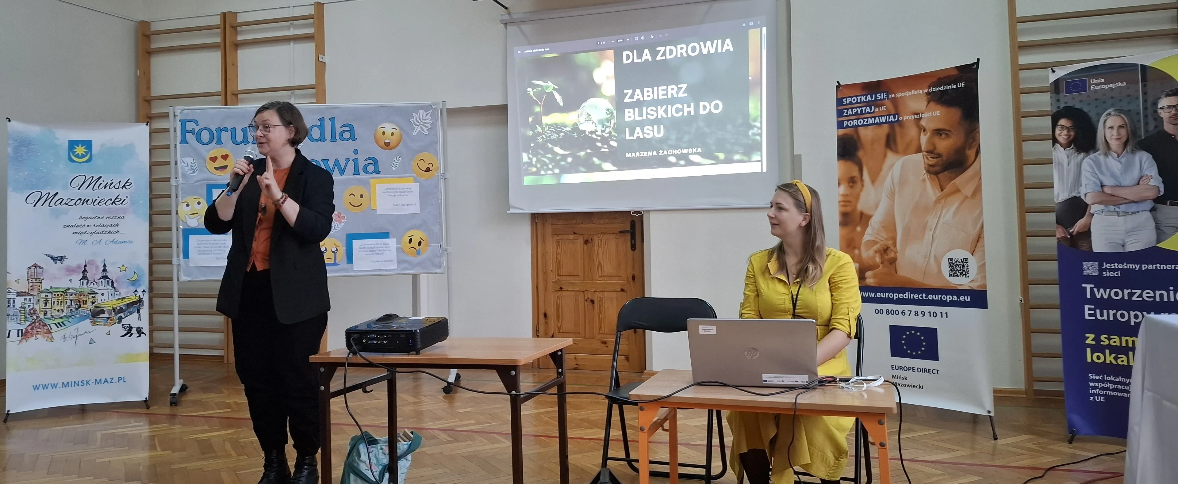 Kobieta w czarnym ubraniu przemawia do mikrofonu w sali z drewnianą podłogą i sprzętem multimedialnym, druga kobieta w żółtej sukience siedzi przy stole z laptopem, za nimi banery promocyjne i ekran z prezentacją.