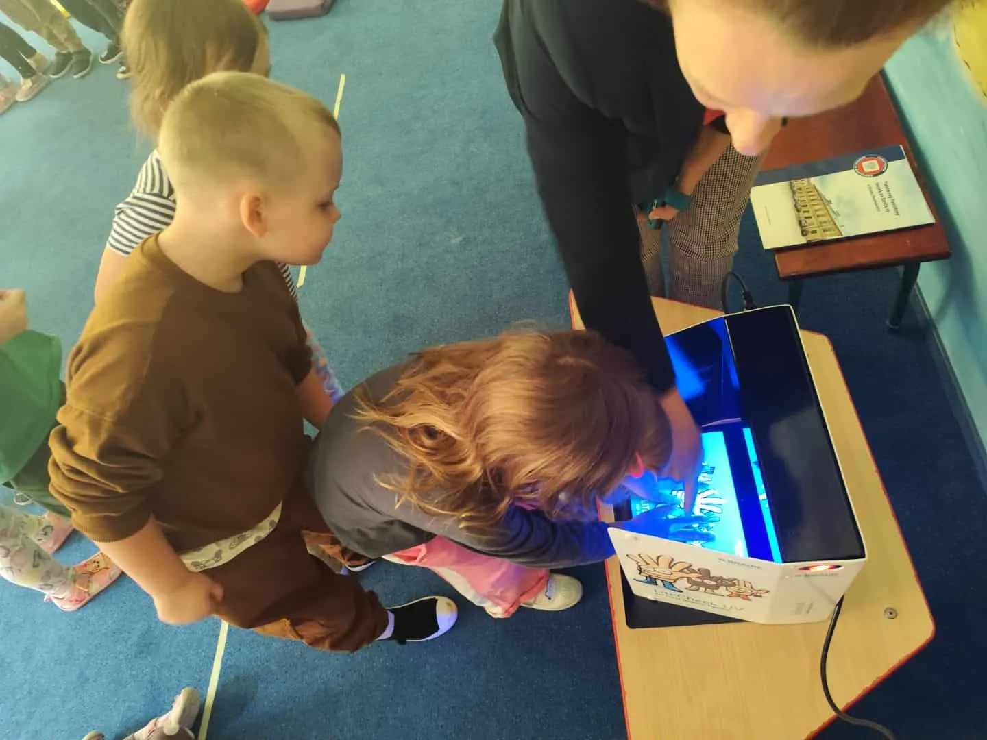 Trzy dzieci i dorosły przy stole z urządzeniem emitującym niebieskie światło, dzieci położone ręce na świetlnym panelu, na podłodze niebieski dywan, obok leży książka.
