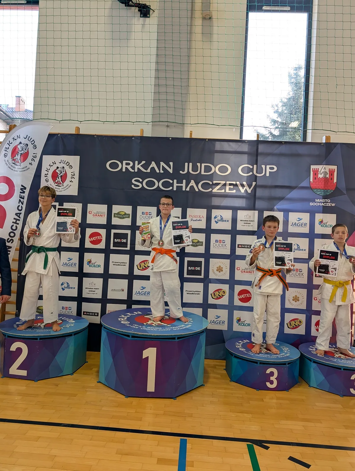 Czterech chłopców w judogach stoi na podium w hali sportowej. Dwóch chłopców na pierwszych dwóch miejscach ma pomarańczowy i zielony pas, dwóch pozostałych posiadają pomarańczowy i żółty pas. Wszyscy trzymają medale i dyplomy. Tło stanowi baner z napisem "Orkan Judo Cup Sochaczew" oraz logotypami sponsorów. Za nimi widać siatkę i duże okna.