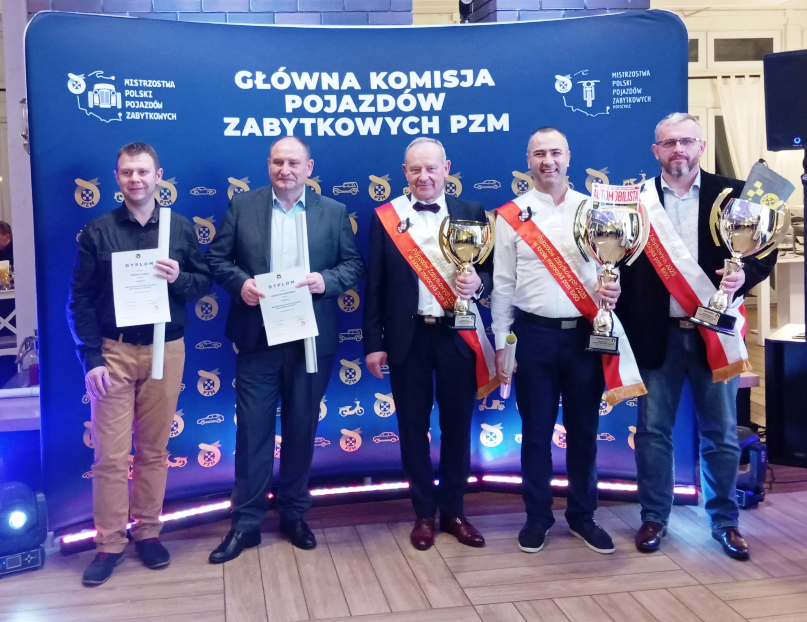 Pięciu mężczyzn stoi na tle niebieskiego baneru z napisem „Główna Komisja Pojazdów Zabytkowych PZM”. Trzech z nich trzyma puchary i ma na sobie czerwone szarfy, dwaj pozostali trzymają dyplomy. Wszyscy ubrani są w półformalne stroje. W tle znajduje się wnętrze budynku z oświetleniem sceny.