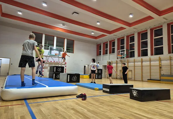 Sześć dzieci ćwiczy na sali gimnastycznej, korzystając z materaca powietrznego i skrzyń do skakania. Sala ma jasne ściany, wysokie okna i drewnianą podłogę, jest dobrze oświetlona. W tle widać piłkochwyty oraz drabinki gimnastyczne.
