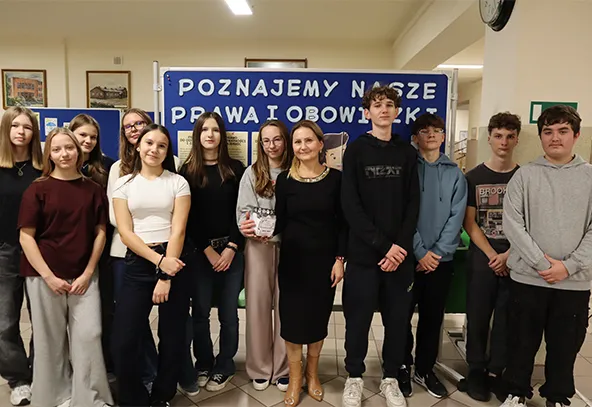 Grupa jedenastu nastolatków i jedna dorosła kobieta stoi w pomieszczeniu szkolnym przed tablicą z napisem „Poznajemy nasze prawa i obowiązki”. Wszyscy patrzą w stronę aparatu.