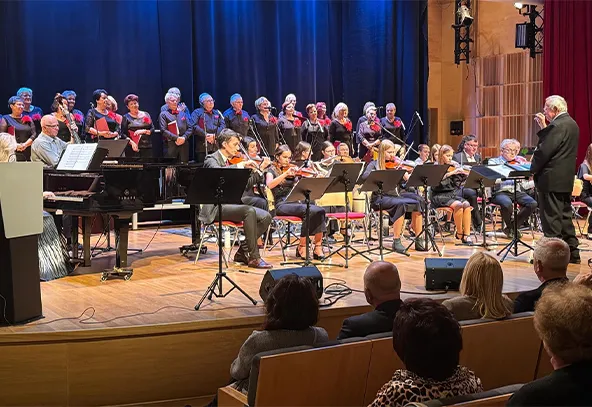 Orkiestra smyczkowa i chór występują na scenie z niebieskim tłem, dyrygent stoi z prawej strony. Publiczność siedzi na widowni w sali koncertowej.