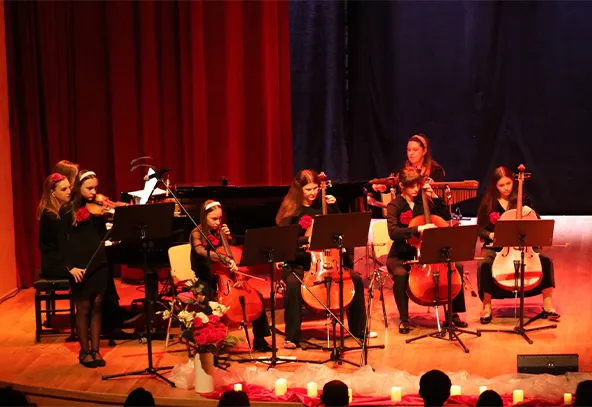 Siedem młodych osób gra na instrumentach smyczkowych na scenie z czerwonym i ciemnoniebieskim tłem. Przed nimi stoją statywy z nutami. Na pierwszym planie widać kwiaty i zapalone świece. W tle stoi fortepian.