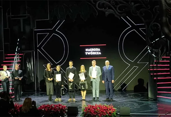 Sześć osób stoi na scenie w rzędzie. Cztery osoby w centrum trzymają dyplomy i torby, obok nich po jednej osobie po każdej stronie. Tło jest czarne z geometrycznymi wzorami i napisem „NAGRODA TWÓRCZA”. Przed sceną znajdują się czerwone kwiaty. Scena jest oświetlona, widownia jest ciemna.