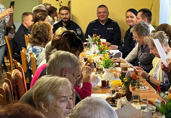 Grupa kilkunastu osób siedzi przy długim stole w pomieszczeniu o żółtych ścianach. Na stole stoją kolorowe kwiaty i napoje. Trzy osoby w mundurach policyjnych siedzą na końcu stołu. Większość osób to starsze kobiety, niektóre czytają dokumenty. Atmosfera przypomina spotkanie lub naradę.