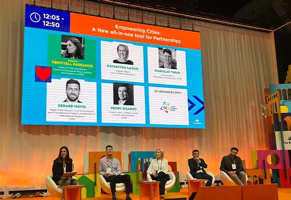 Pięć osób siedzi na scenie w białych fotelach na tle jasnych kolorów i dużego ekranu. Na ekranie są zdjęcia i nazwiska pięciu mówców oraz tytuł sesji "Empowering Cities: A New all-in-one tool for Partnerships". Scena jest oświetlona ciepłym światłem, a tło ma kolor jasnopomarańczowy i beżowy.