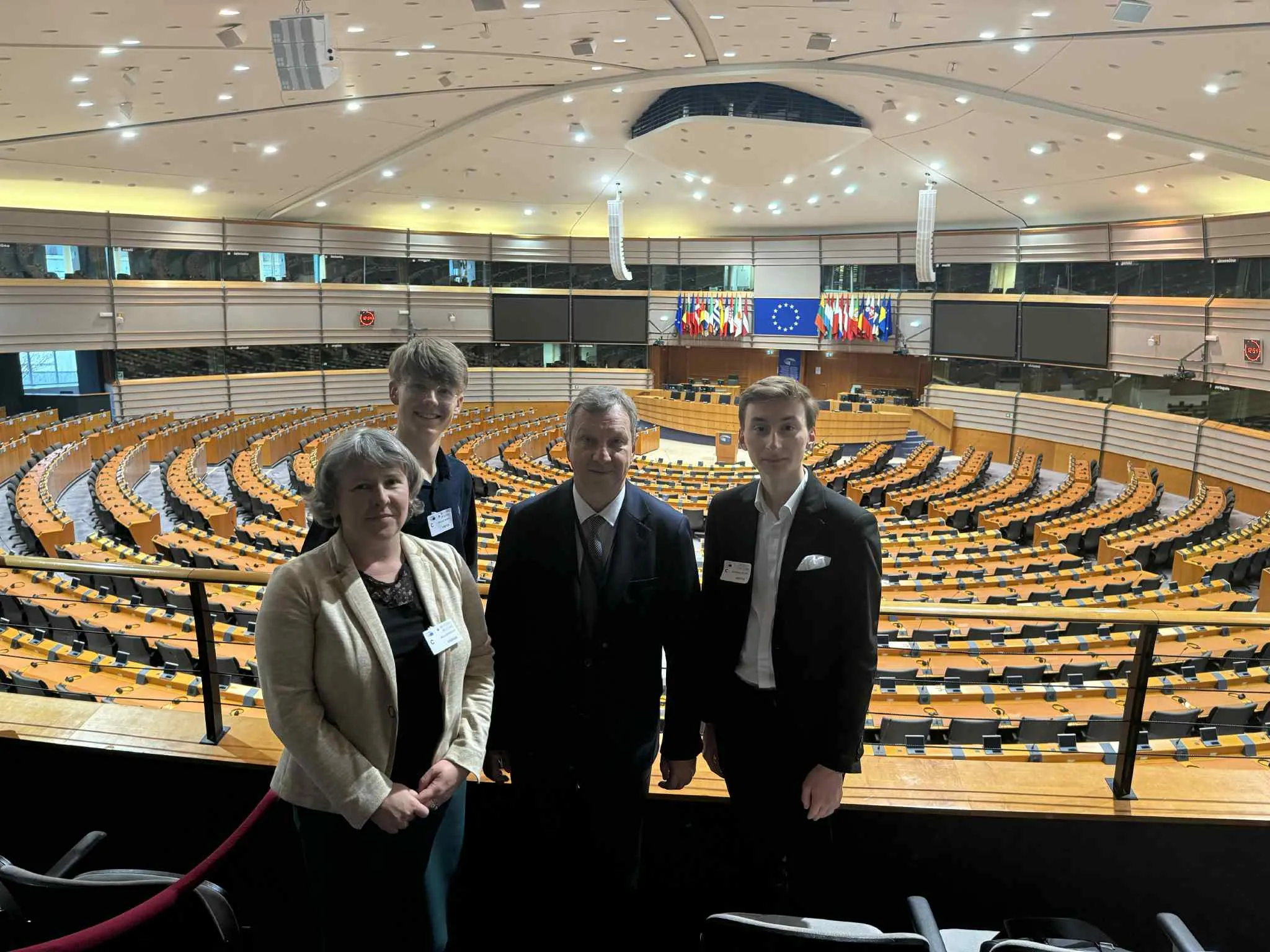 Cztery osoby stoją na balkonie sali konferencyjnej z dużą liczbą pustych miejsc siedzących ułożonych w półokręgi, w tle widoczne flagi państw członkowskich Unii Europejskiej i niebieska flaga UE.