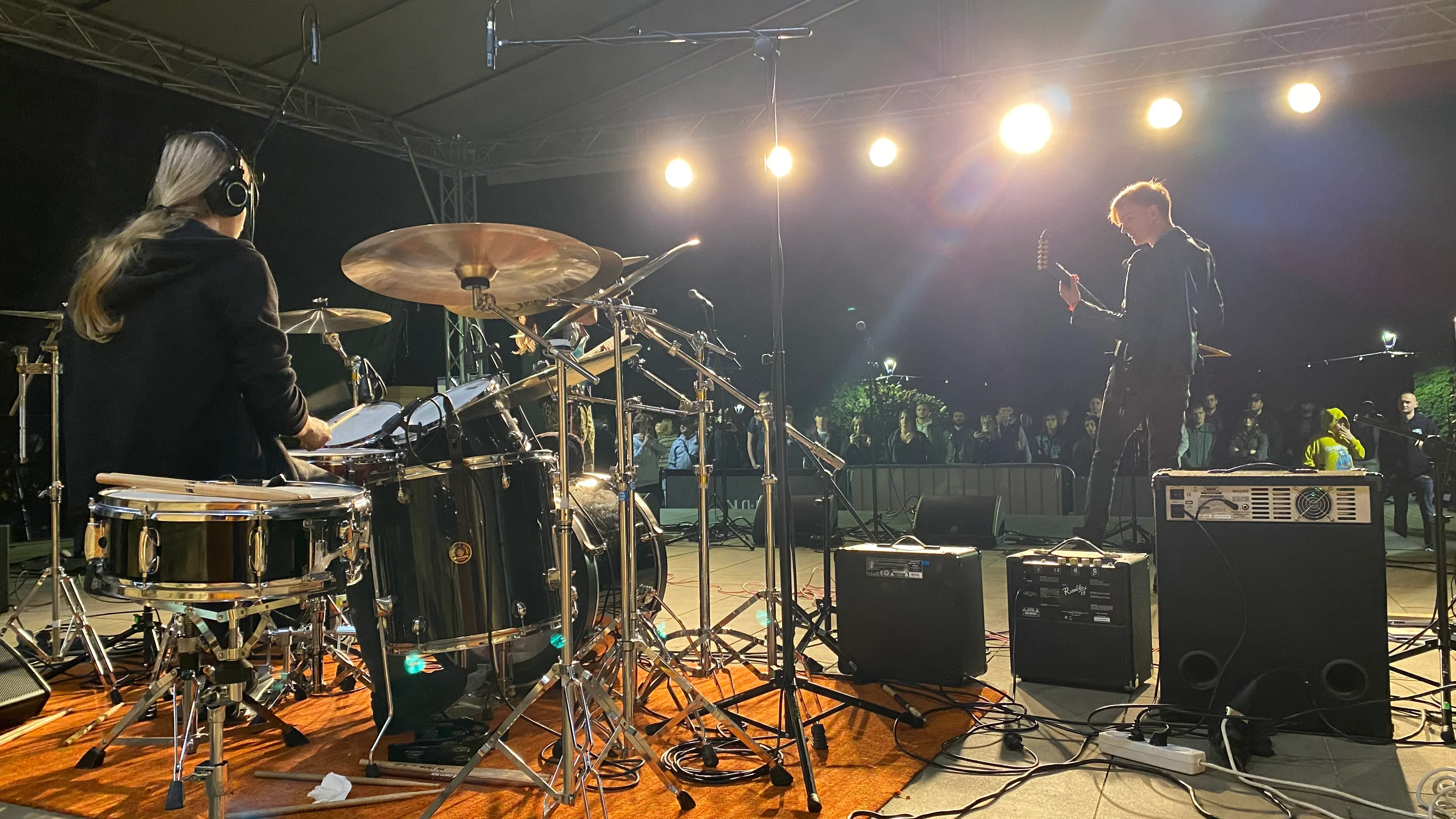 Perkusista i gitarzysta grają na scenie na zewnątrz nocą, oświetleni jasnymi reflektorami, przed nimi stoi publiczność za barierką.