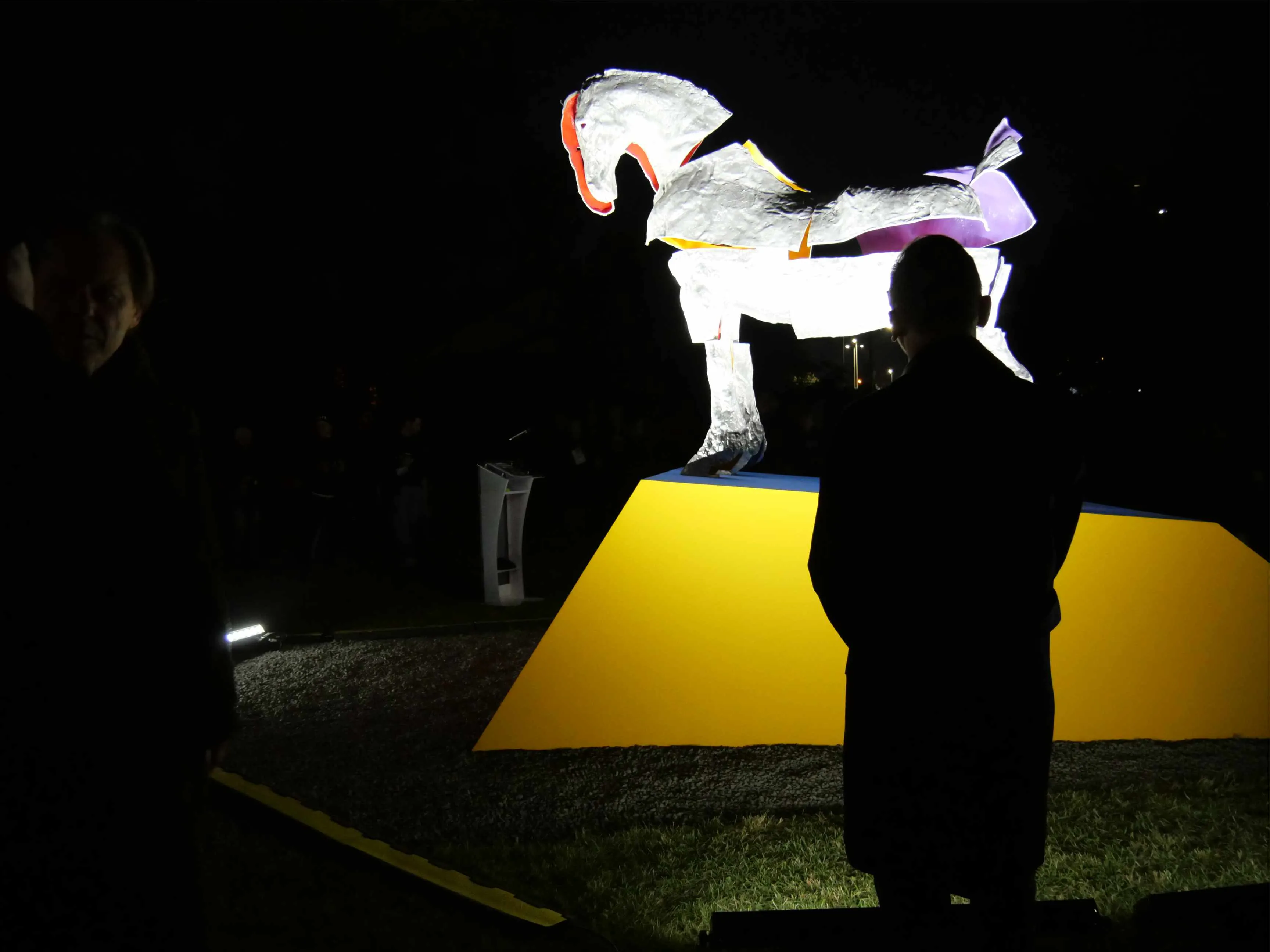 Rzeźba konia na żółtym cokole oświetlona nocą, obok stoi sylwetka osoby, w tle lekko widoczna druga osoba i ciemne otoczenie.