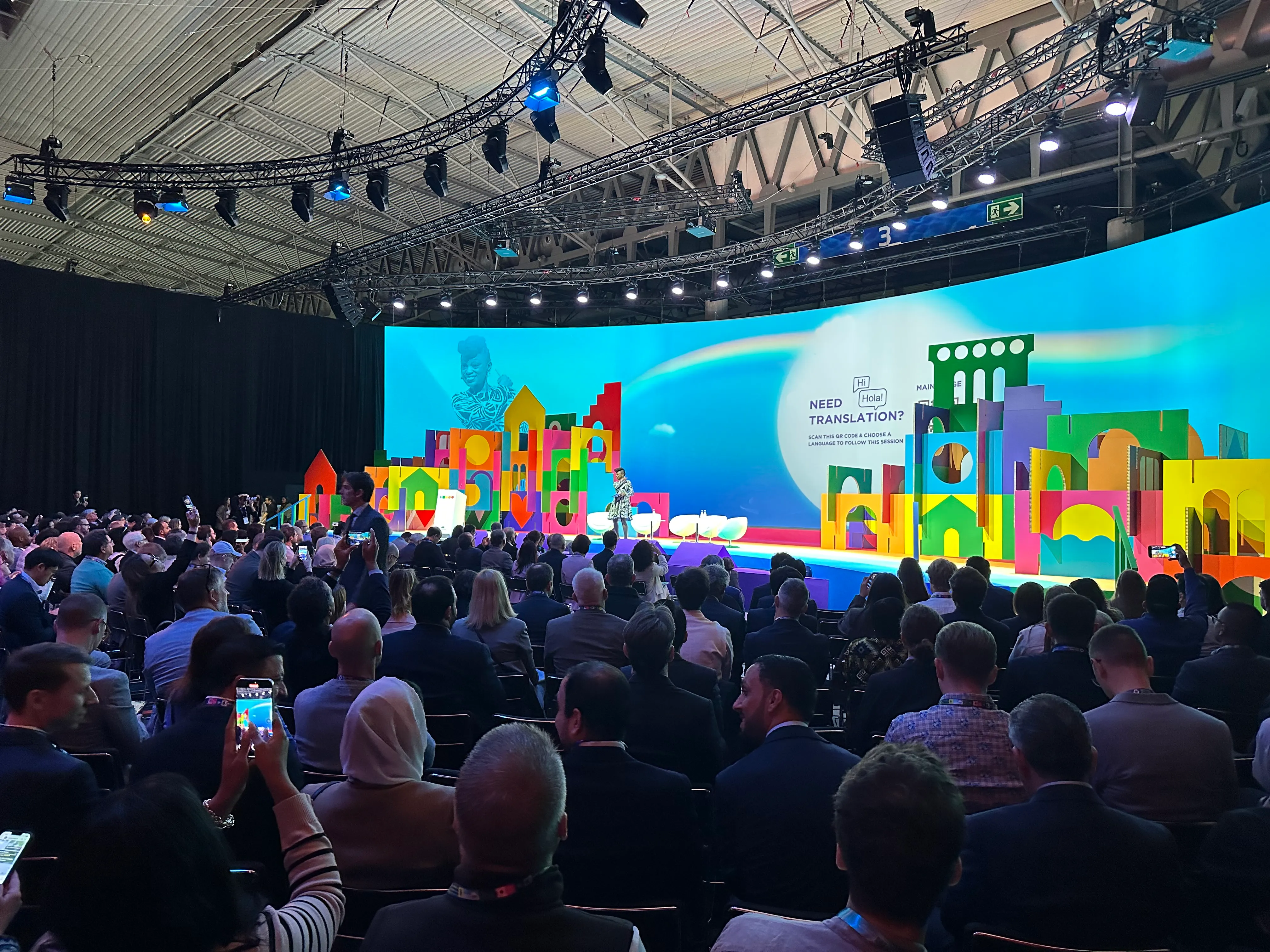 Wnętrze sali konferencyjnej z dużą liczbą siedzących osób zwróconych ku kolorowej scenie z prezentacją i kobietą przemawiającą. Na tle jasnoniebieskim wielokolorowe dekoracje sceniczne. Widoczne oświetlenie i elementy konstrukcyjne sufitu.