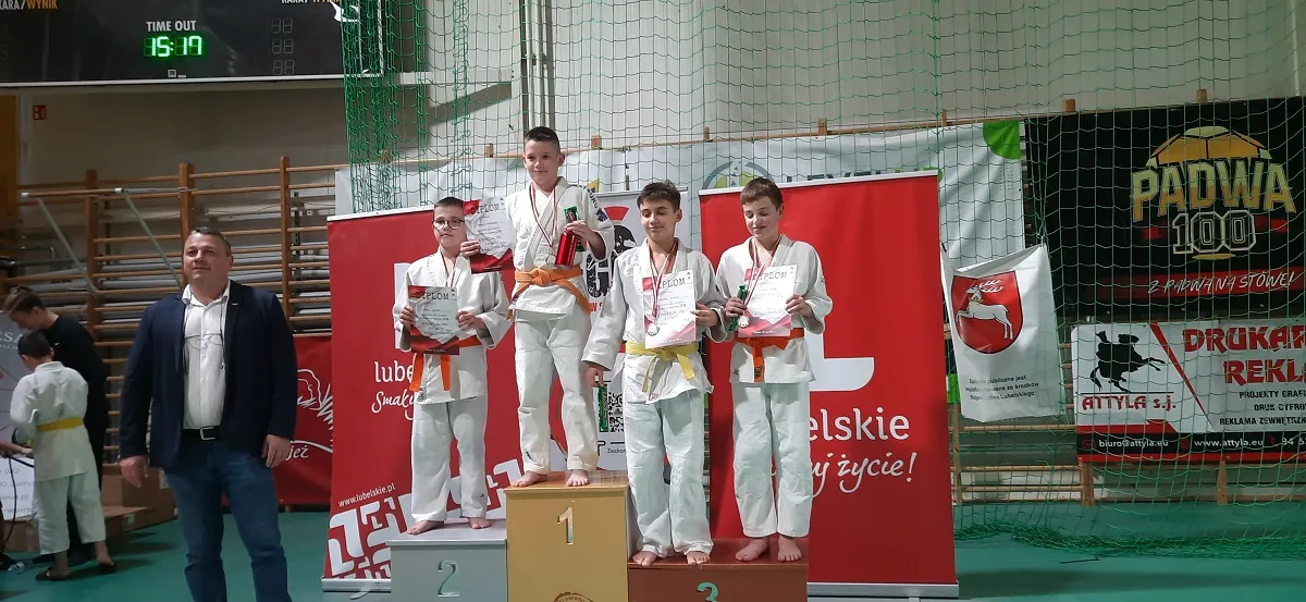 Czterech chłopców w judogach stoi na podium z medalami i dyplomami na hali sportowej, po lewej stoi mężczyzna w marynarce, w tle siatka i banery reklamowe.