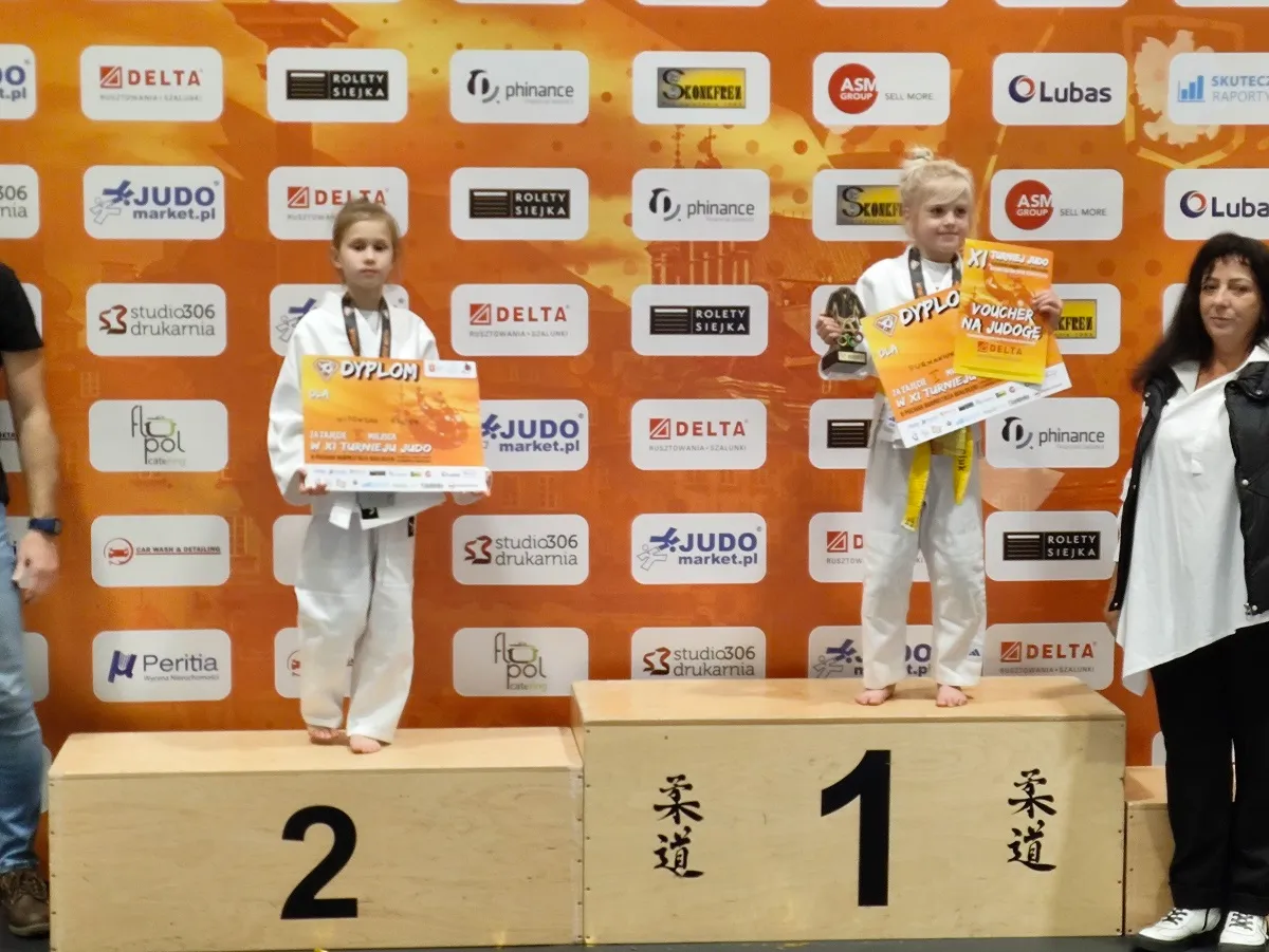 Dwie dziewczynki w białych judogach stoją na podium na miejscach pierwszym i drugim. Dziewczynka na pierwszym miejscu trzyma puchar i dyplom, ma żółty pas. Dziewczynka na drugim miejscu trzyma dyplom, ma biały pas. W tle pomarańczowa ścianka z logo sponsorów. Po prawej stoi kobieta w białej koszuli i czarnej kamizelce. Po lewej częściowo widoczna osoba dorosła.