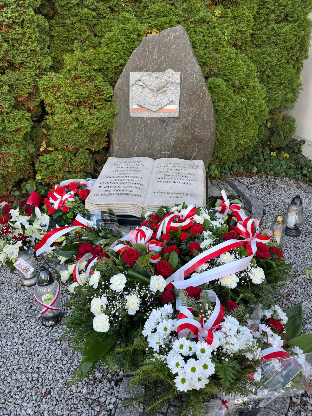 Pomnik z kamienia z zegarem słonecznym i otwartą książką z inskrypcją, otoczony zielonymi krzewami. Przed pomnikiem liczne wiązanki kwiatów w barwach biało-czerwonych i znicze na szarym żwirze.